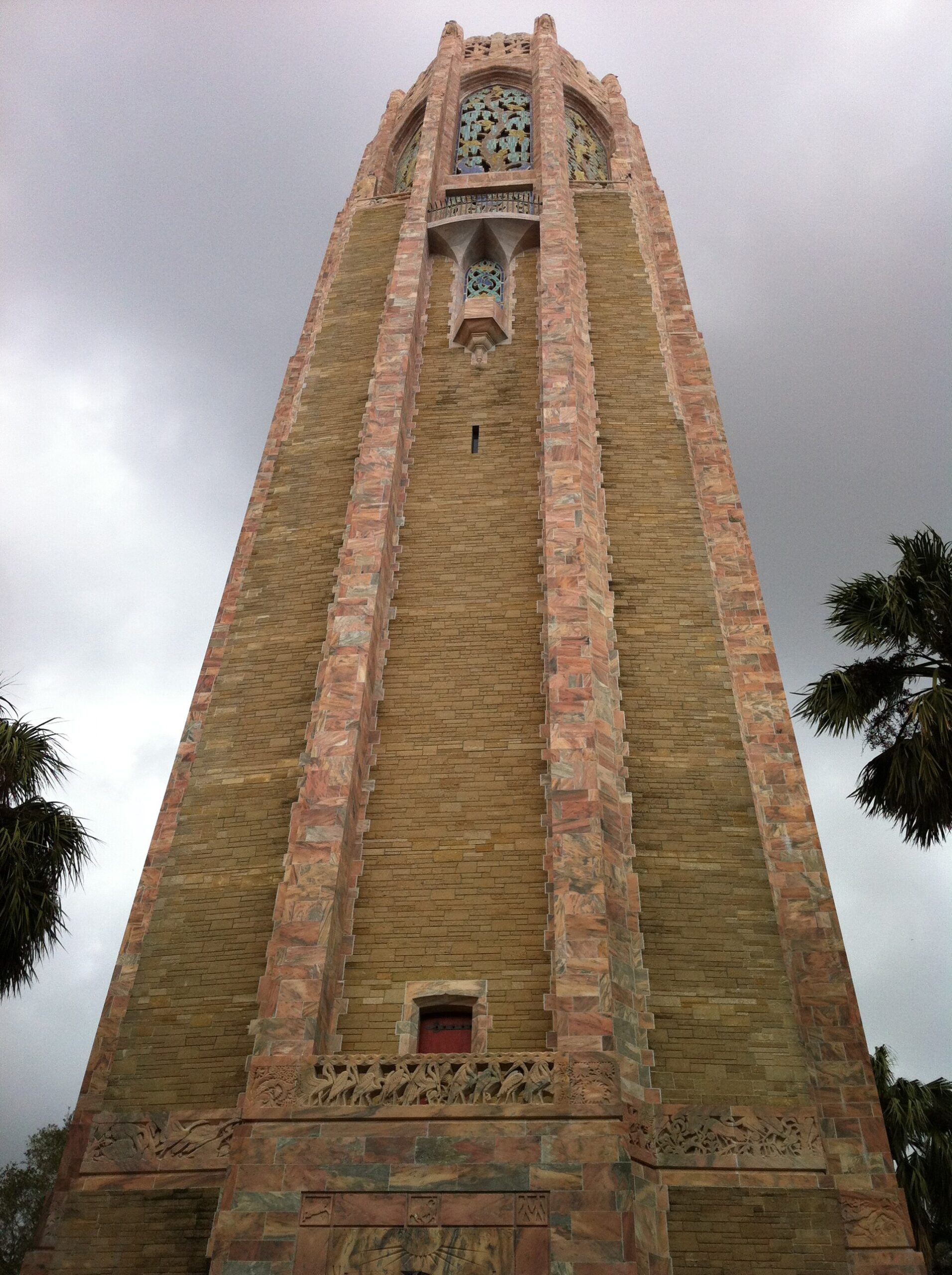 The tall pink marble and coquina Bok Tower at Lake Wales, Florida, seen from below against a grey sky, with carved stone reliefs around its base and stained glass in its upper window