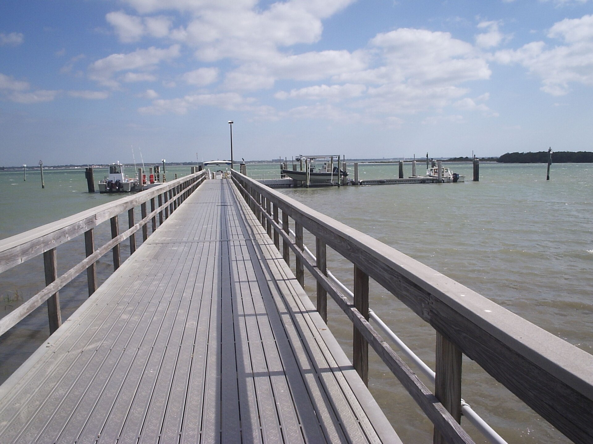 A long wooden boardwalk pier running out over calm green water toward boat slips and moored boats, with a low wooded island on the horizon