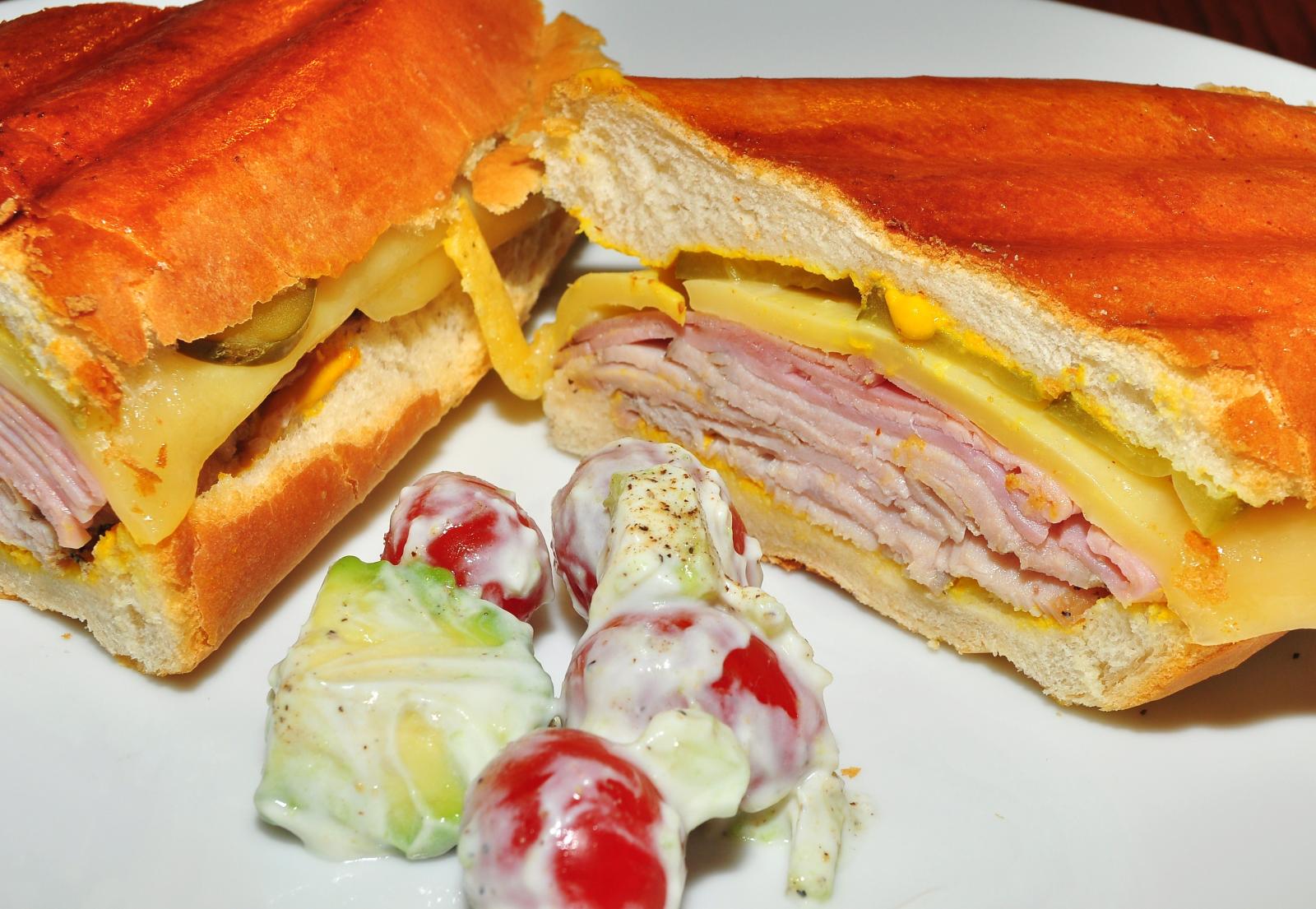 A pressed Cuban sandwich cut in half on a white plate, showing layers of ham, roast pork, Swiss cheese, mustard and pickle inside a golden pressed crust