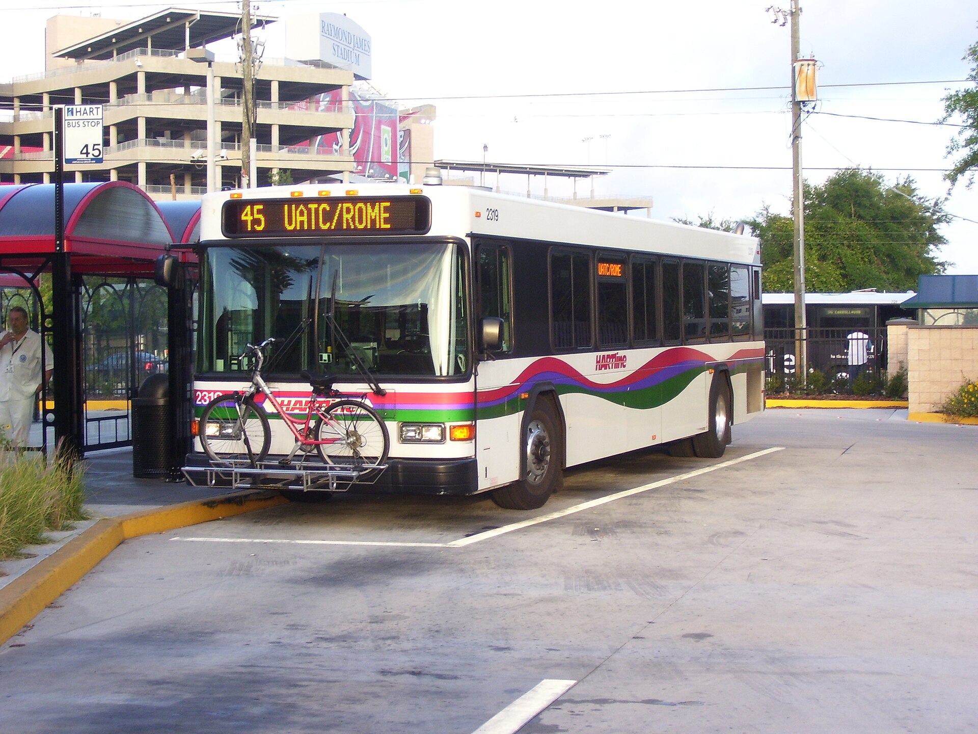 A white HART bus with colored striping at a covered bus stop, its destination sign lit, with a bicycle on the front rack and Raymond James Stadium behind