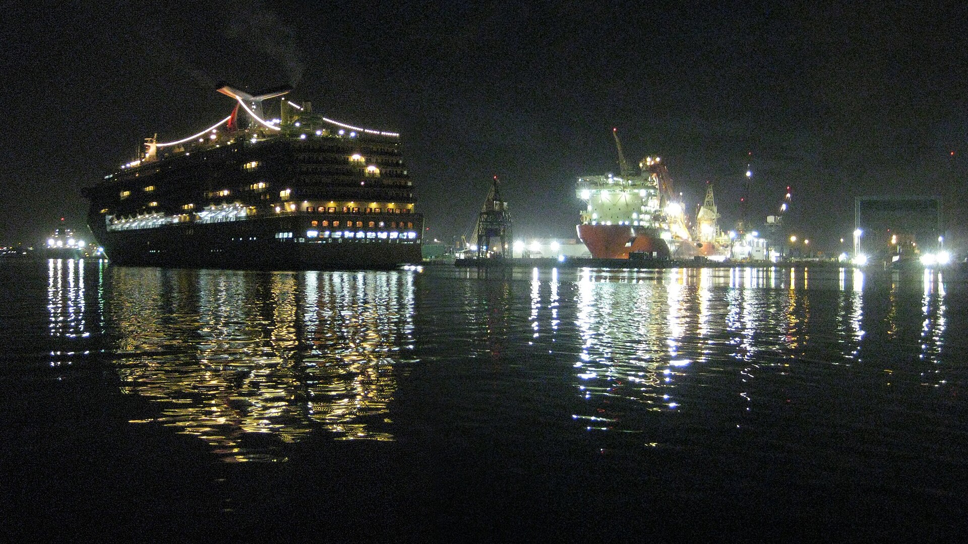 A cruise ship lit up entering a port channel in darkness before dawn, with shipyard cranes and a vessel in drydock on the far bank and their lights reflected in still water