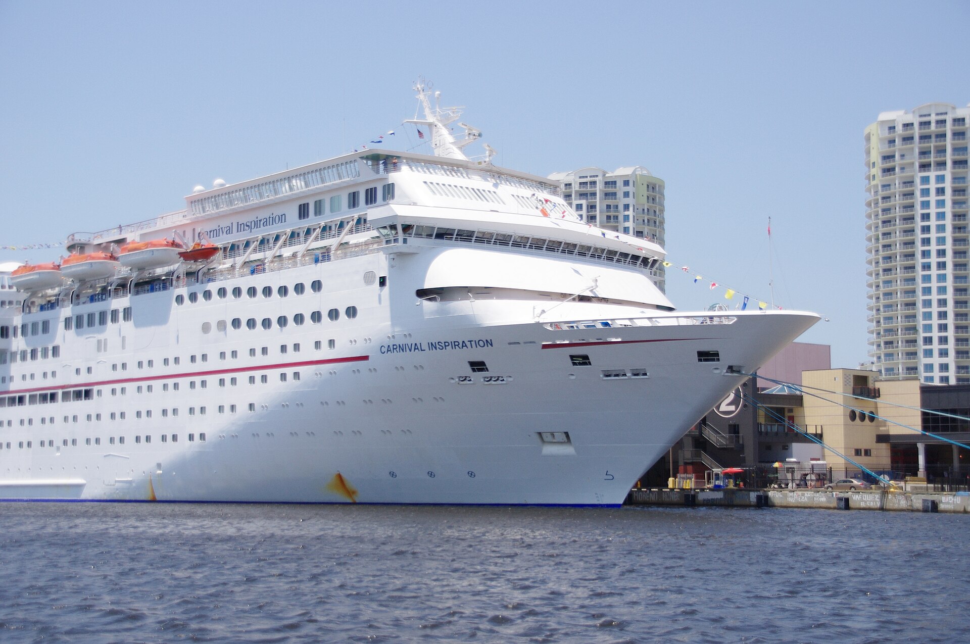 A white midsize cruise ship moored bow-on alongside a Tampa cruise terminal building marked with a large number 2, with Channelside high-rise condominiums behind