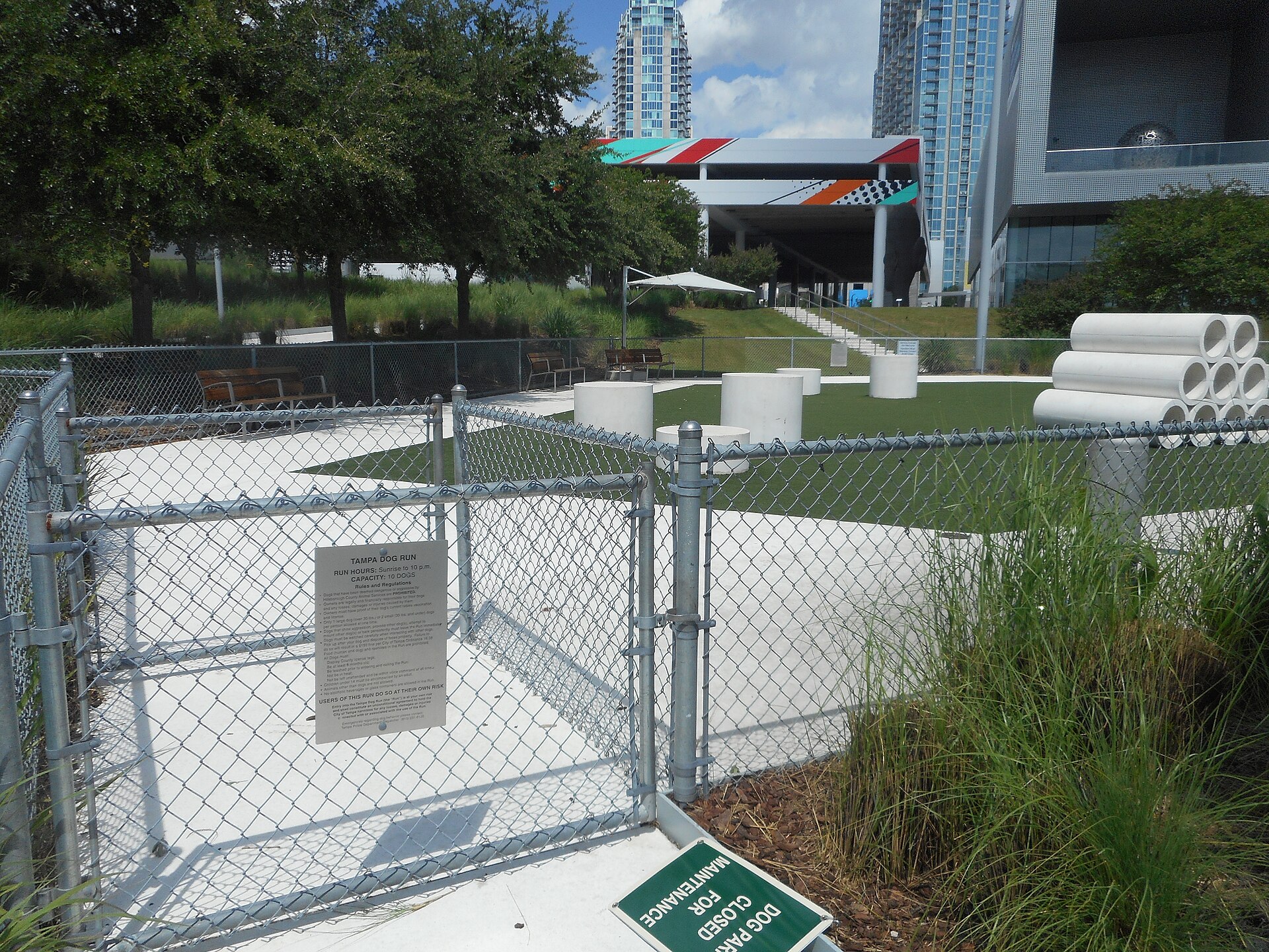A fenced dog run beside the Tampa Riverwalk at Curtis Hixon Waterfront Park, with a posted rules sign, artificial turf and downtown high-rises behind