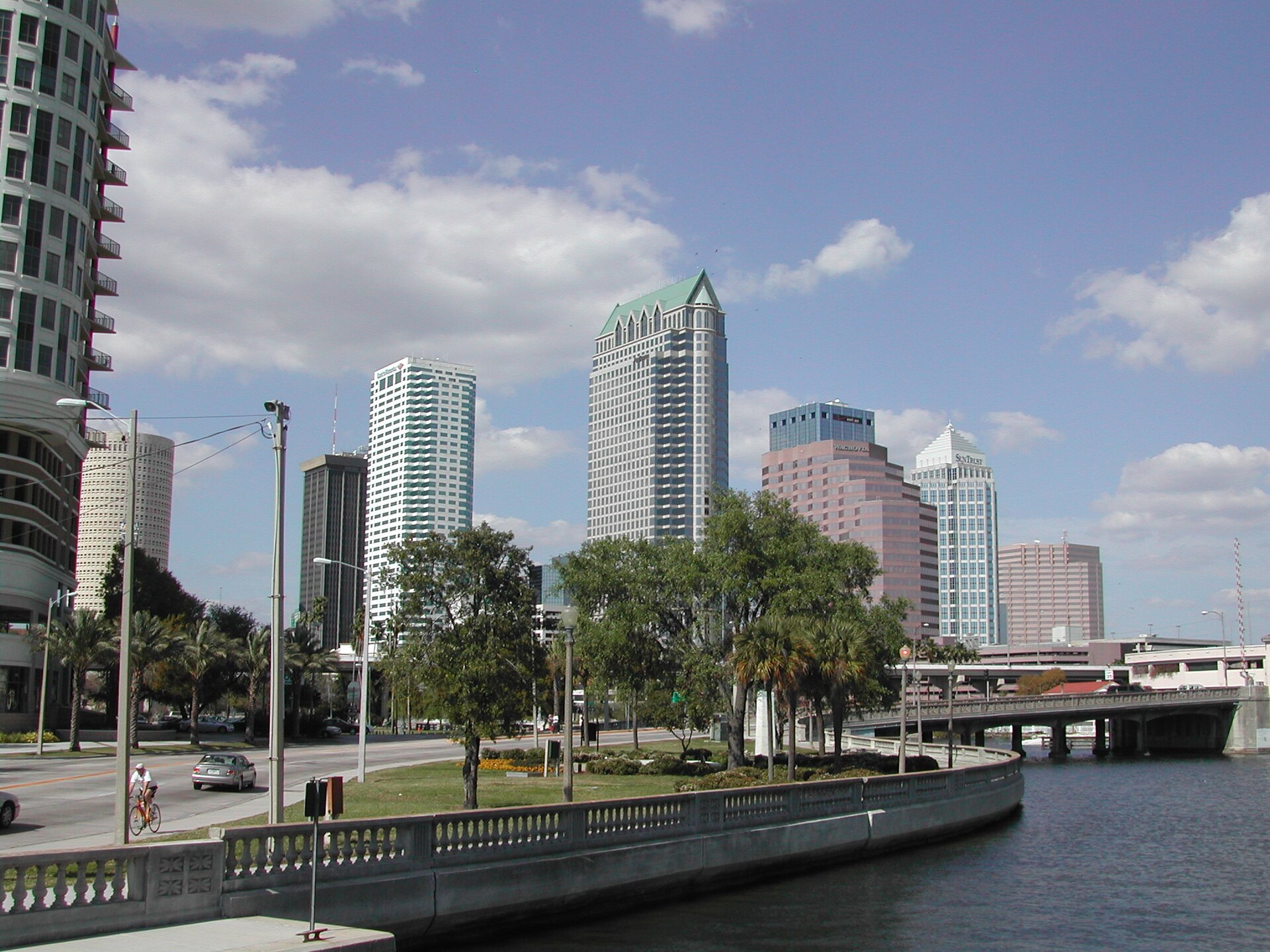 The downtown Tampa skyline of glass and stone towers seen across parkland from the balustraded sidewalk at the northern end of Bayshore Boulevard, under fair-weather cumulus