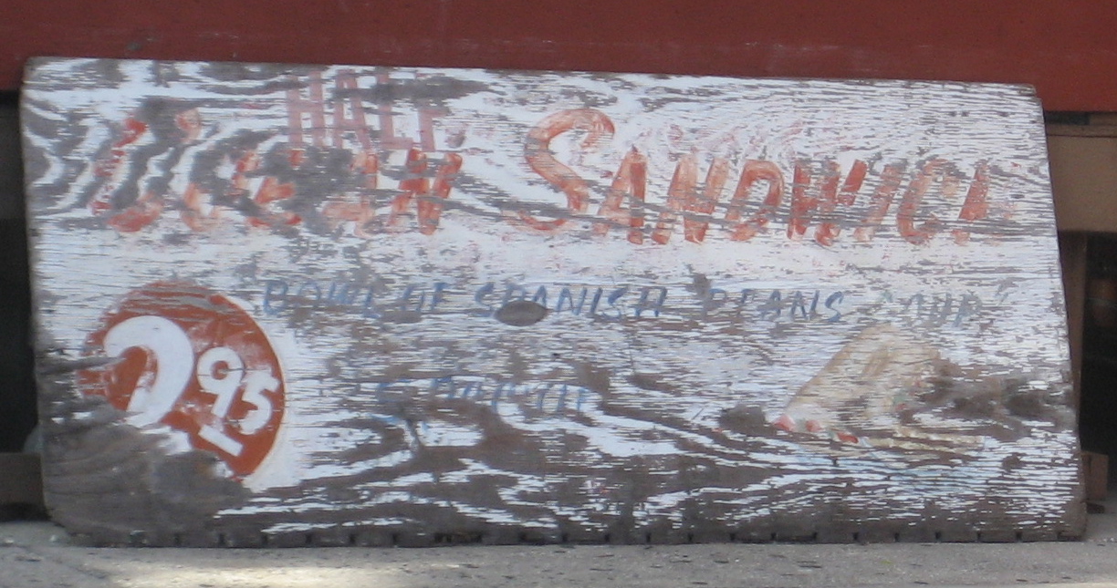A weathered painted wooden sign leaning against a wall in Ybor City reading Half Cuban Sandwich, bowl of Spanish bean soup, two dollars ninety-five