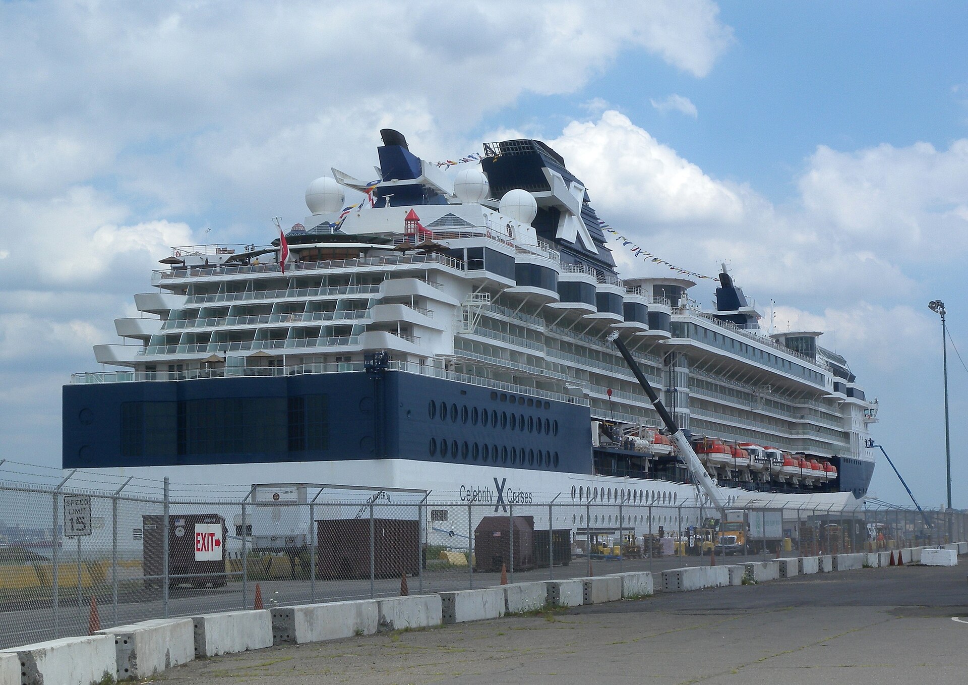 Celebrity Cruises' Celebrity Summit docked at a pier, with its navy-blue stern and stacked balconies behind a chain-link fence and a 15 mph speed-limit sign