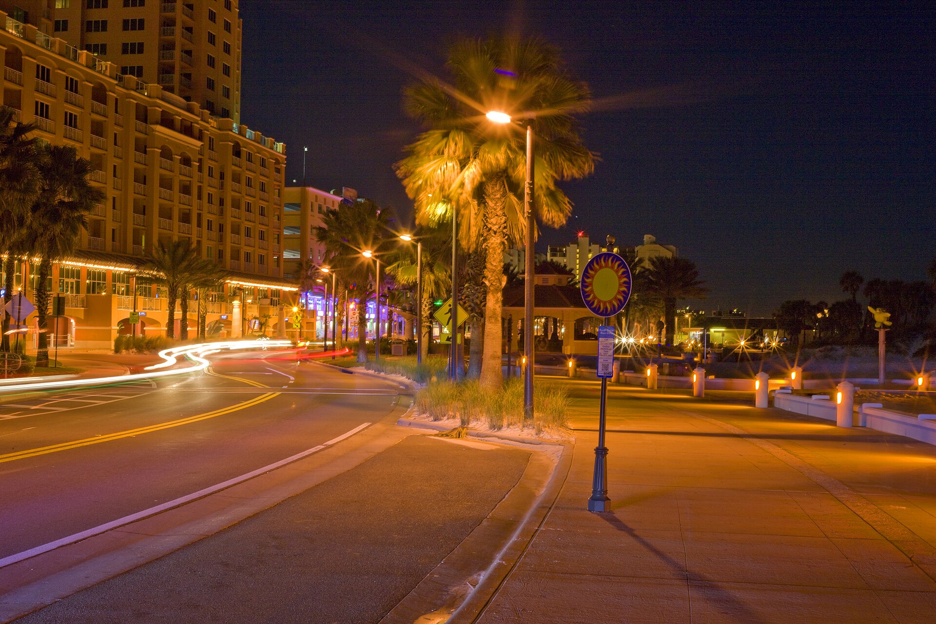 A beachfront road on Clearwater Beach at night, with a large hotel tower on the left, palm trees and streetlights, a wide lit promenade on the right and light trails from passing cars