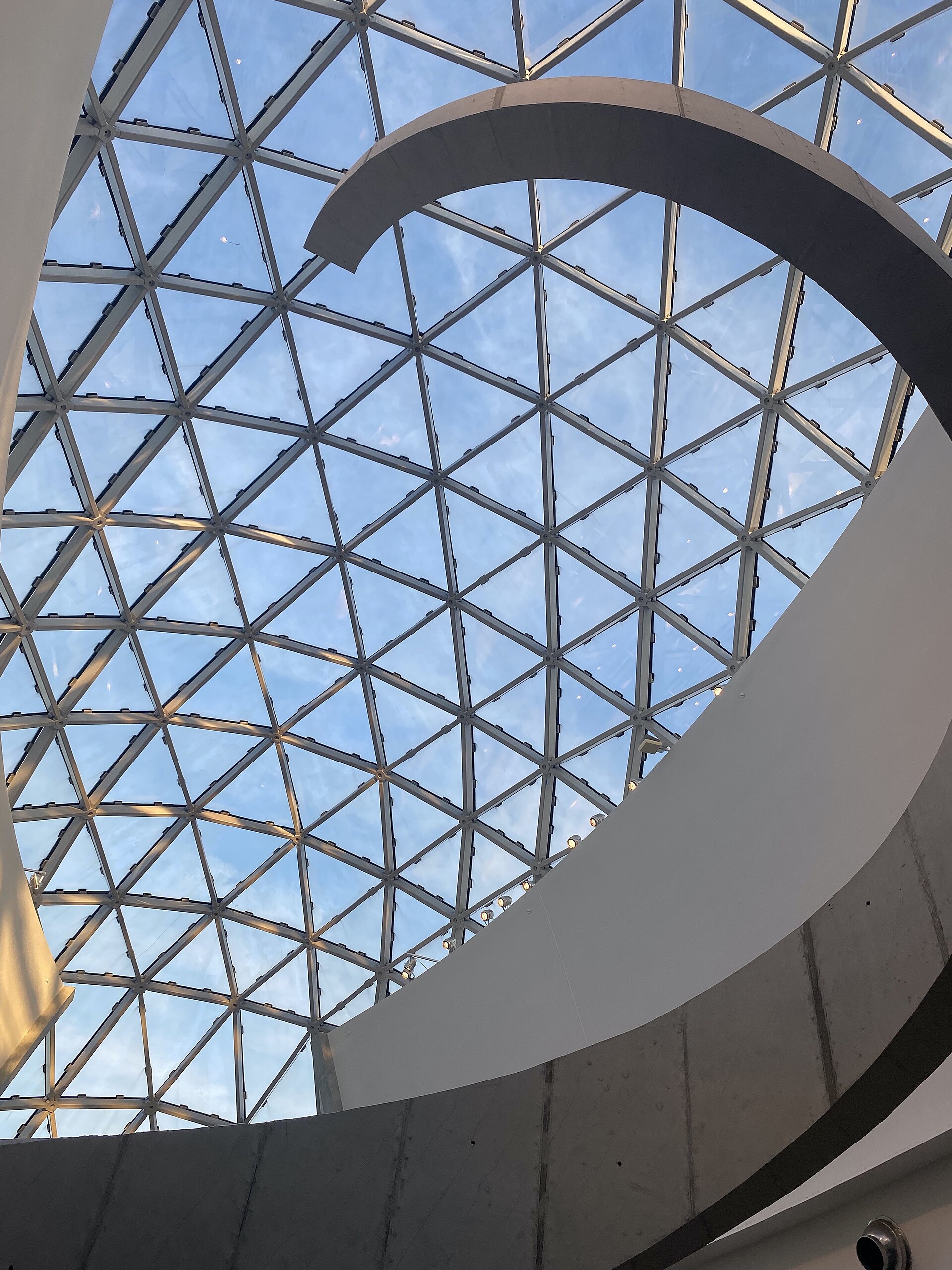 Looking up the helical concrete staircase inside the Dalí Museum toward the geodesic glass ceiling of triangular panels, with blue sky beyond