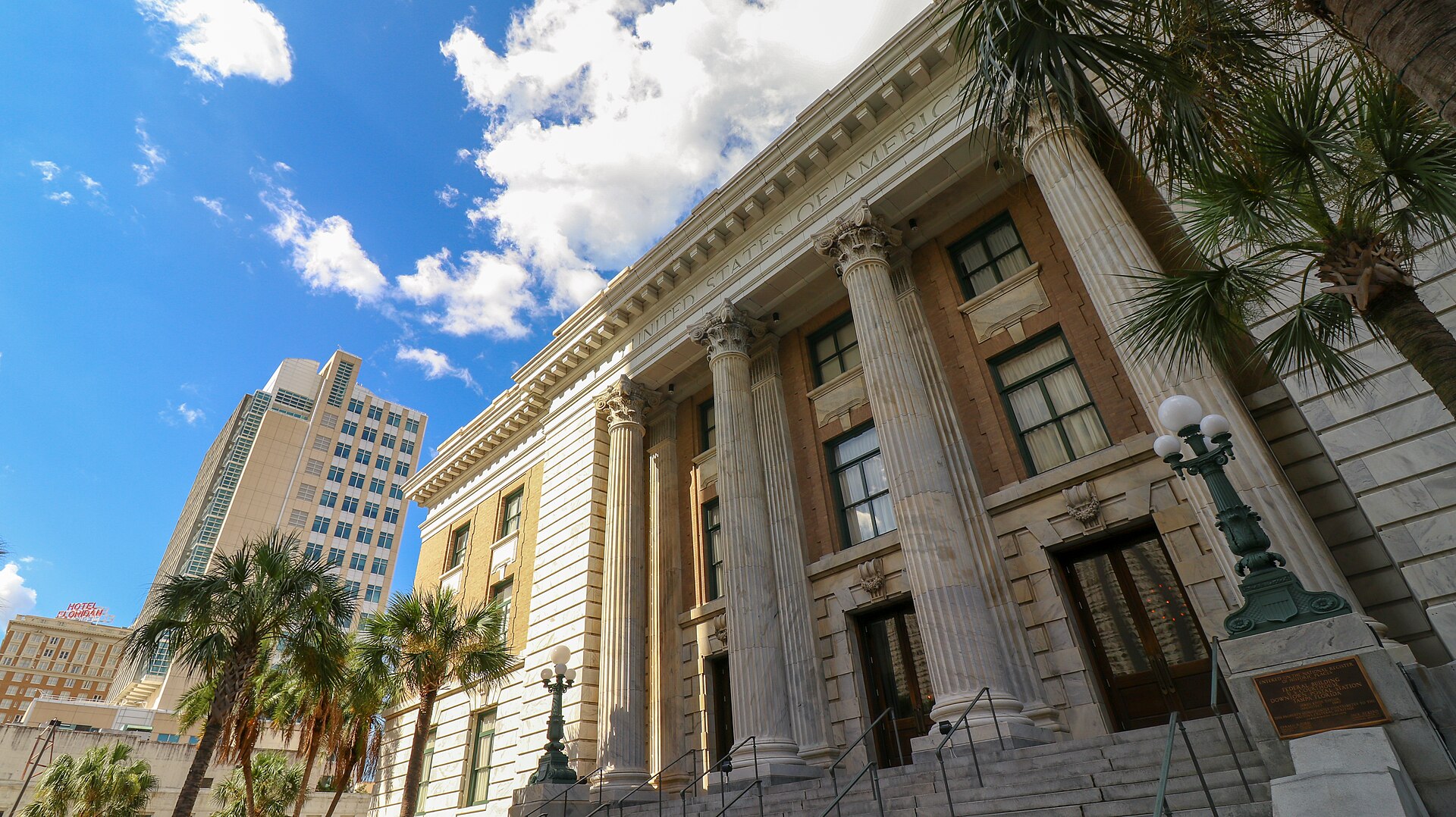 The columned marble facade of the former United States courthouse in downtown Tampa, now the Le Méridien hotel, with palm trees in front and a modern tower and the Hotel Floridan sign behind
