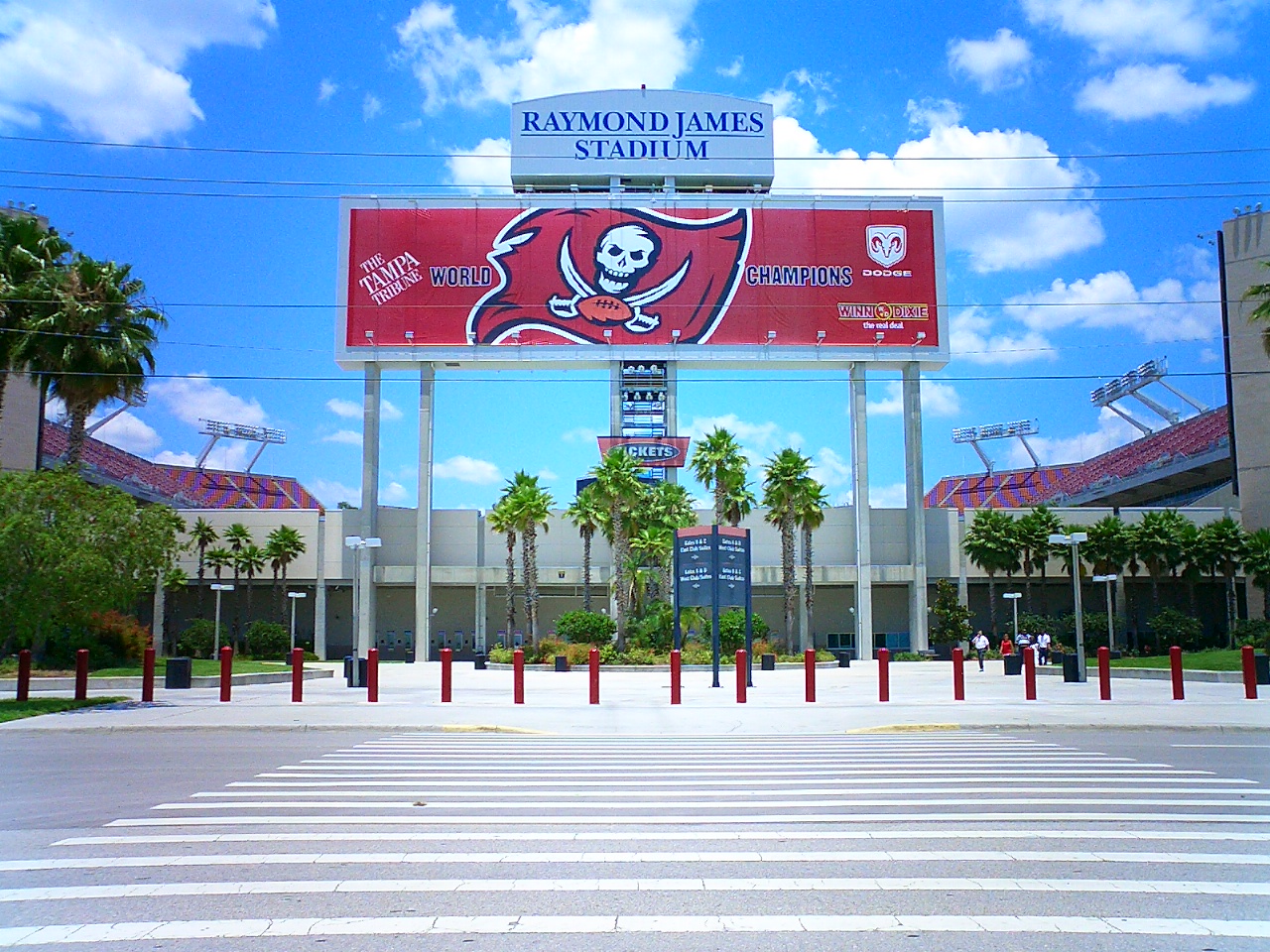 The Raymond James Stadium entrance plaza seen across a wide crosswalk, with a large Buccaneers banner under the stadium name sign and palm trees in front of the gates