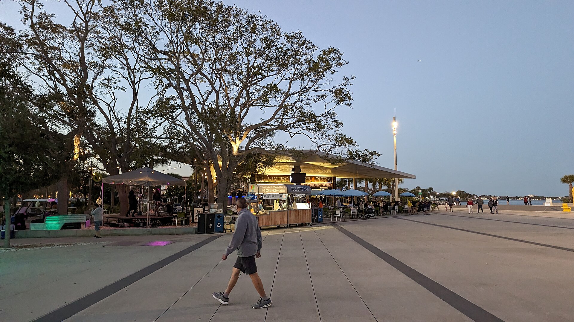 Spa Beach Bistro on the St. Pete Pier at dusk, with a lit food counter selling pizza and ice cream under a large tree, string lights and blue umbrellas over outdoor tables