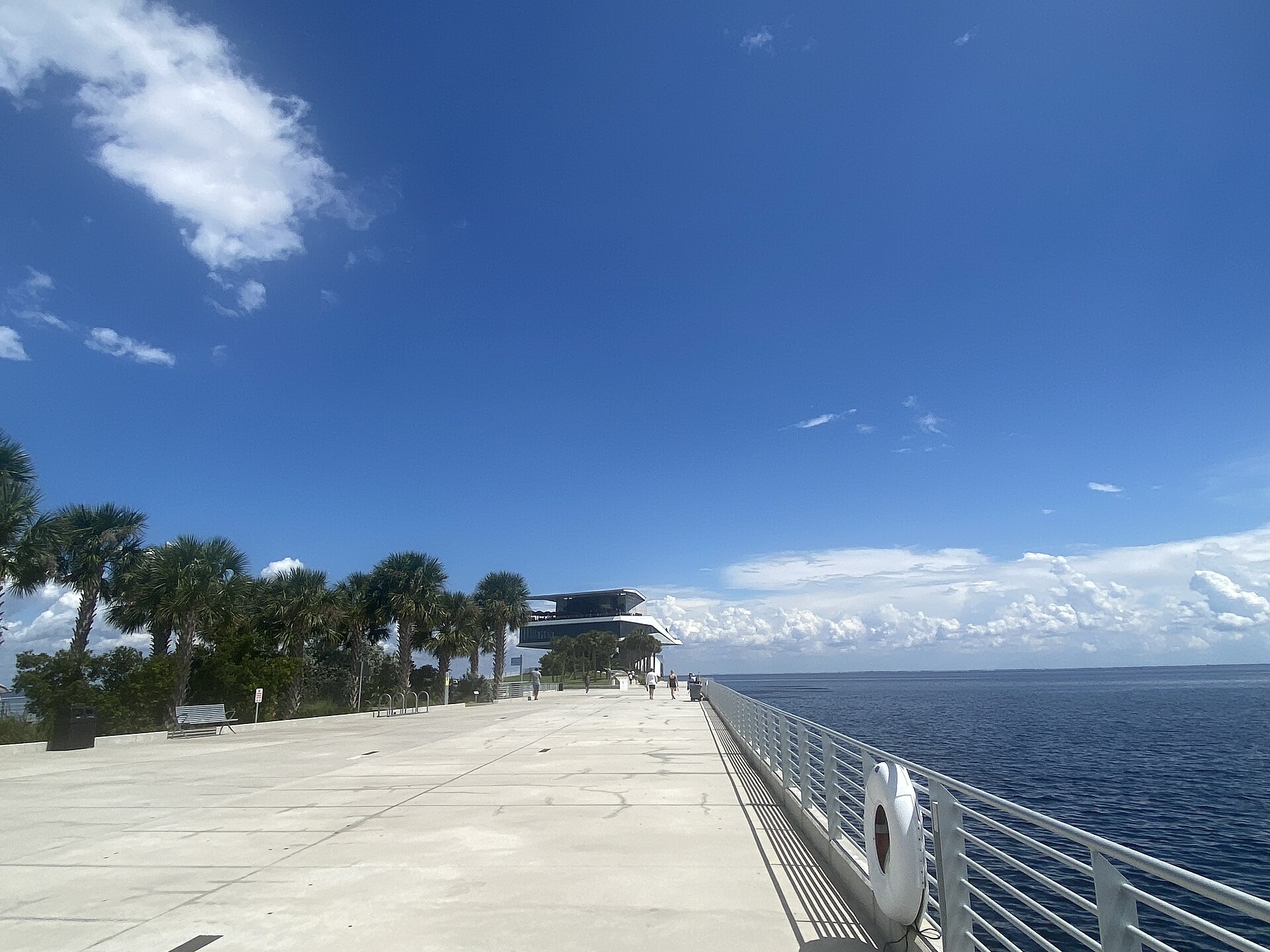 The wide concrete deck of the St. Pete Pier running out over blue water toward the Pier Point building, with palms along one side and a life ring on the railing