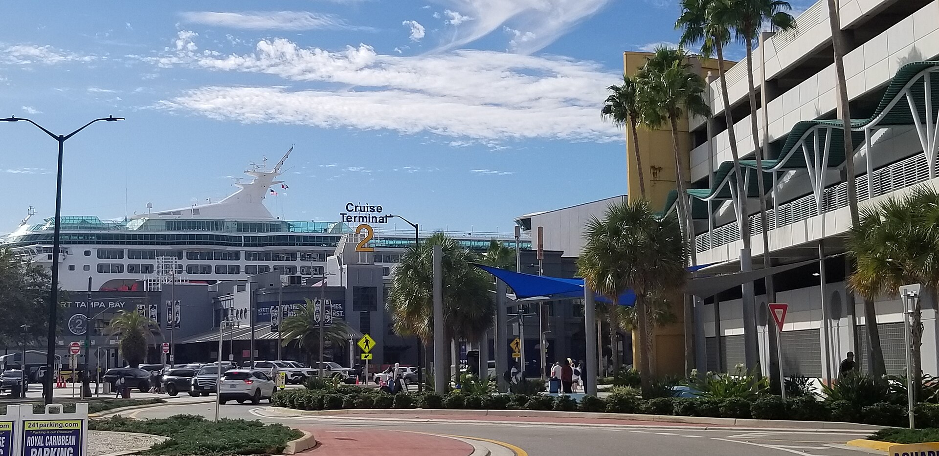 Port Tampa Bay's Cruise Terminal 2 seen from the Channelside Drive roundabout, with a Royal Caribbean ship behind the terminal, passengers with luggage on the sidewalk and the port parking garage on the right